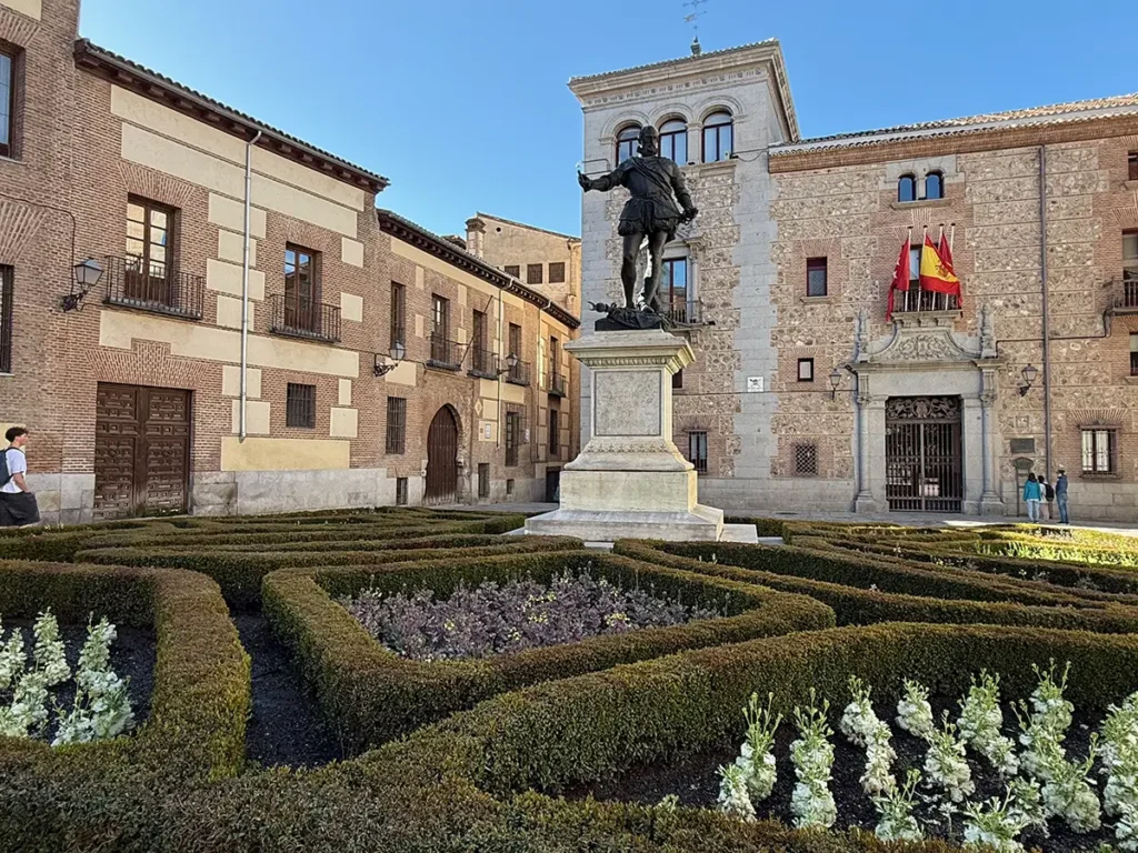 Statue in the Plaza de la Villa in Madrid, Spain, set within neatly trimmed formal gardens and surrounded by historic brick and stone buildings beneath a clear blue sky