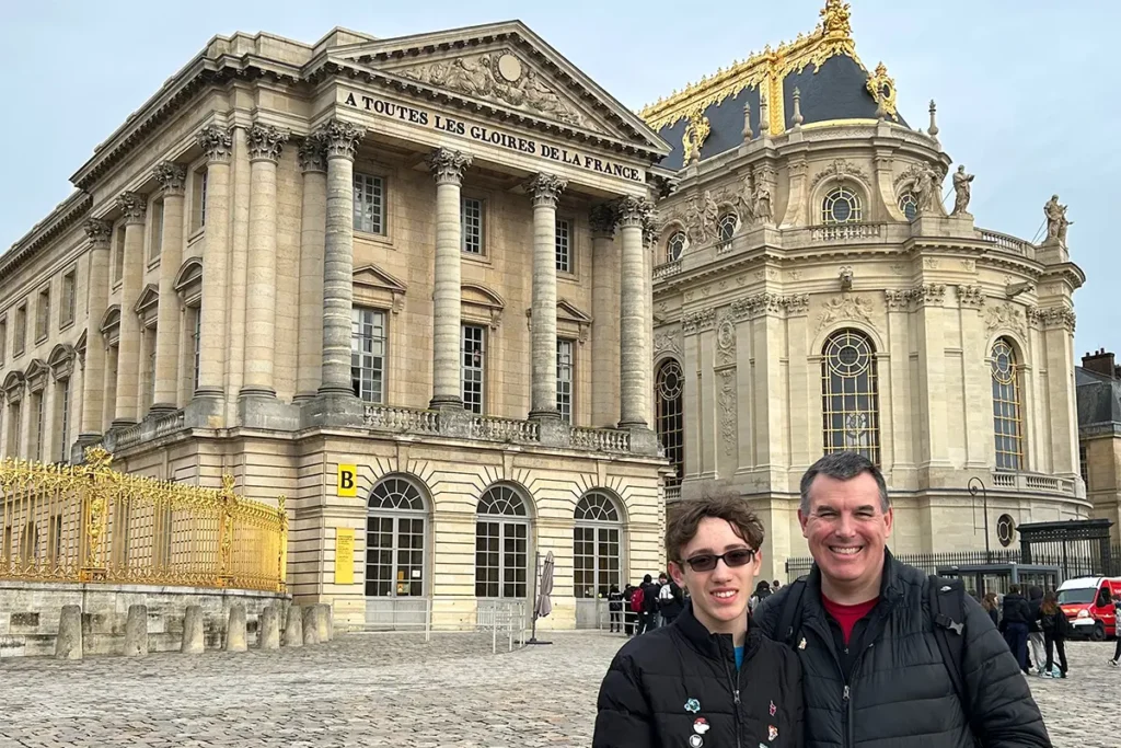 Father and son smile at the Palace of Versailles before the Gabriel Wing, inscribed 'A TOUTES LES GLOIRES DE LA FRANCE,' and the ornate Royal Chapel.