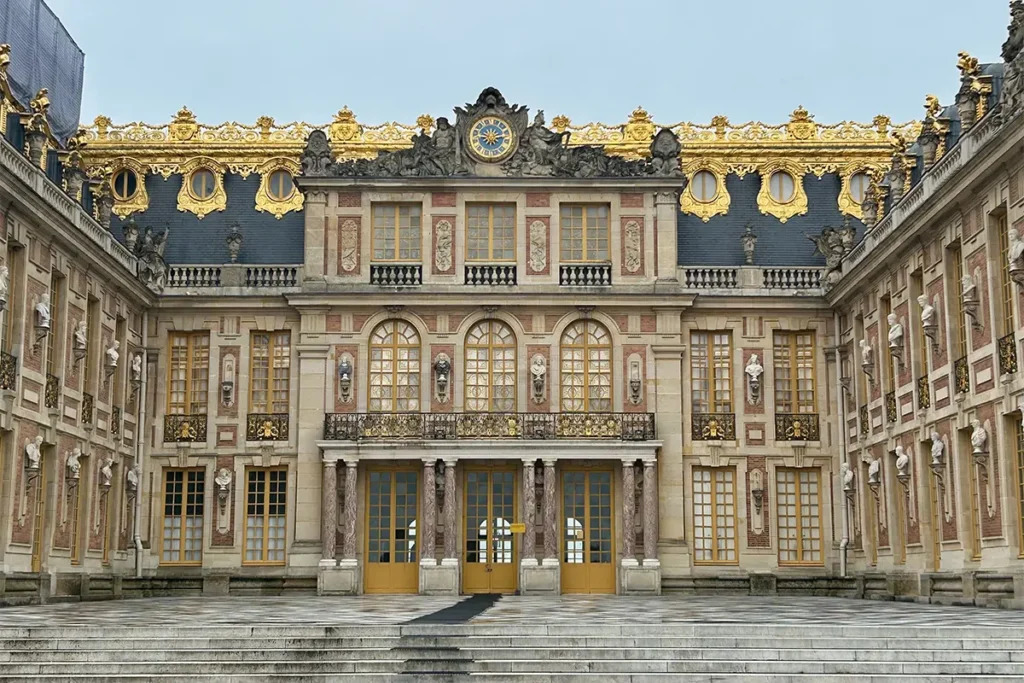 The Royal Court of Versailles featuring a brick facade, white marble busts, golden window frames, and an ornate clock atop the central balcony.