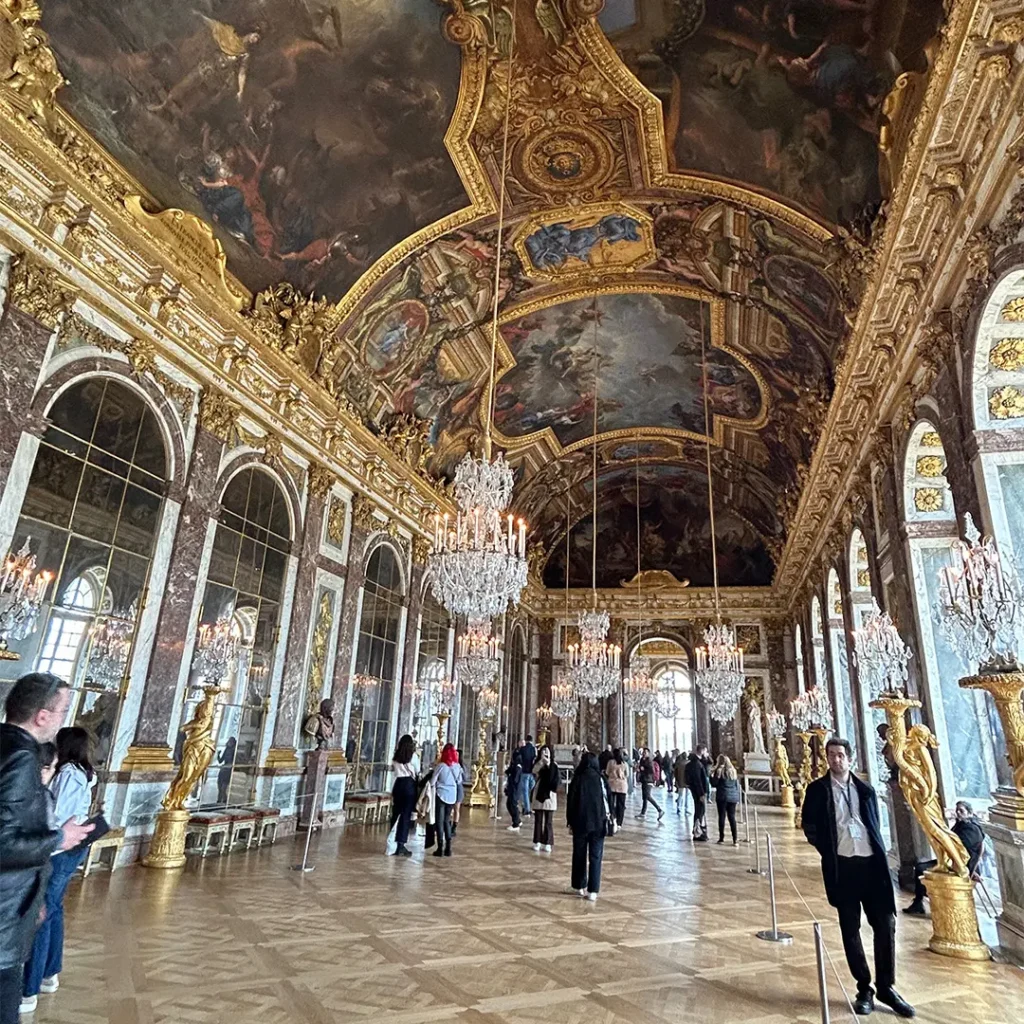 The Hall of Mirrors at Versailles, featuring high arched mirrors, ornate gold ceilings with frescoes, crystal chandeliers, and a parquet floor.
