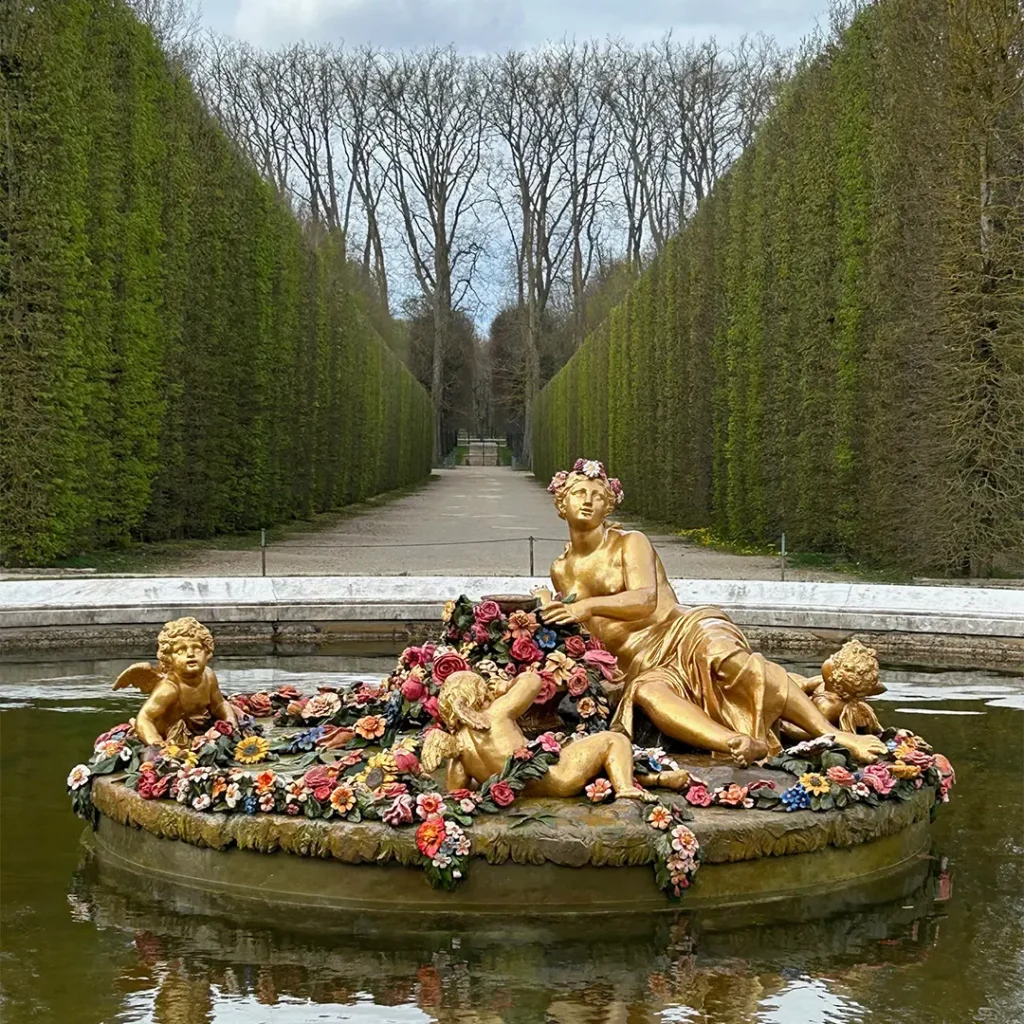 Golden statue of Flora and cherubs adorned with colorful flowers in a fountain, set against a long, hedge-lined path in the Versailles gardens.