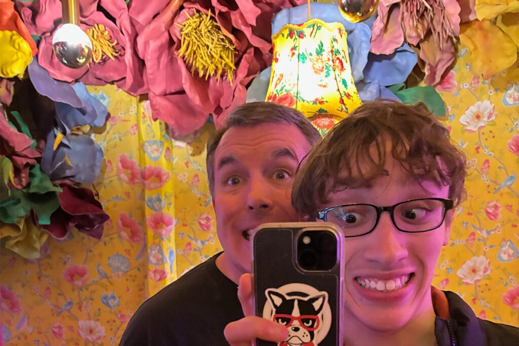 Mirror selfie of two men making silly faces at Rosi La Loca in Madrid, surrounded by giant colorful paper flowers and vibrant floral wallpaper.