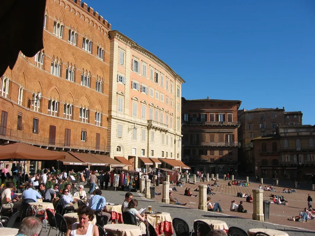 Outdoor cafés and people relaxing in Piazza del Campo in Siena, Italy, with historic brick and pastel-colored buildings surrounding the sloped square under a clear blue sky