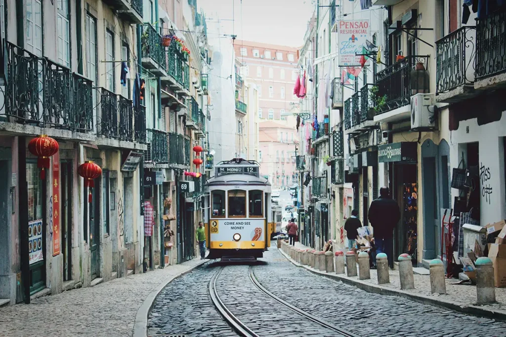 Yellow Lisbon tram traveling along narrow cobblestone tracks between tall, closely packed buildings with wrought-iron balconies, shopfronts, and hanging lanterns in a historic neighborhood