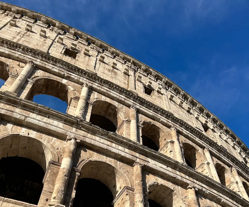 Upward view of the Colosseum in Rome, showing weathered stone arches and columns against a bright blue sky
