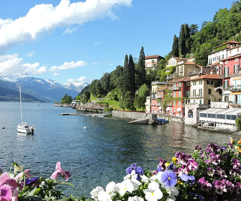 Colorful flowers in the foreground frame a view of Lake Como, featuring the charming village of Varenna with its tiered buildings and a lone sailboat