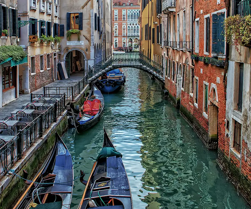 A narrow Venetian canal lined with historic brick buildings and docked gondolas, featuring a small arched bridge and an empty waterside cafe terrace