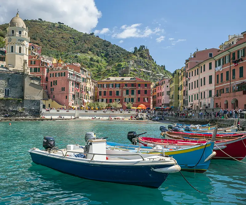 Several colorful motorboats bob in the turquoise harbor of Vernazza, Italy, with vibrant pink and yellow buildings and a church tower along the shore