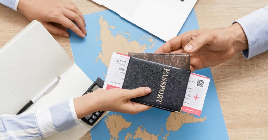 Travel advisor with client, exchanging a passport and boarding pass over a desk with a world map, notebook, calculator, and laptop