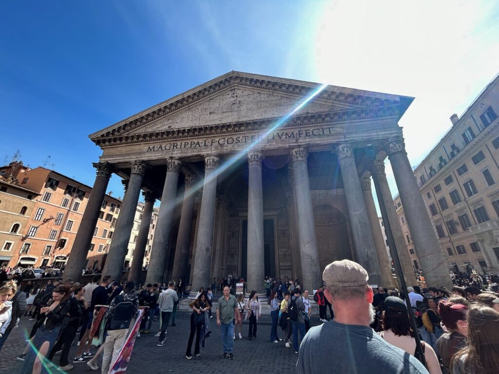 Photo of the Pantheon in Rome, showcasing its massive Corinthian columns and the Latin inscription on the pediment under a bright, clear sky. A large crowd of tourists gathers in the foreground in the Piazza della Rotonda.