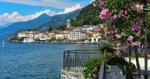 Pink flowers bloom along a stone railing, overlooking the blue waters of Lake Como and a picturesque town with mountain peaks in the background.