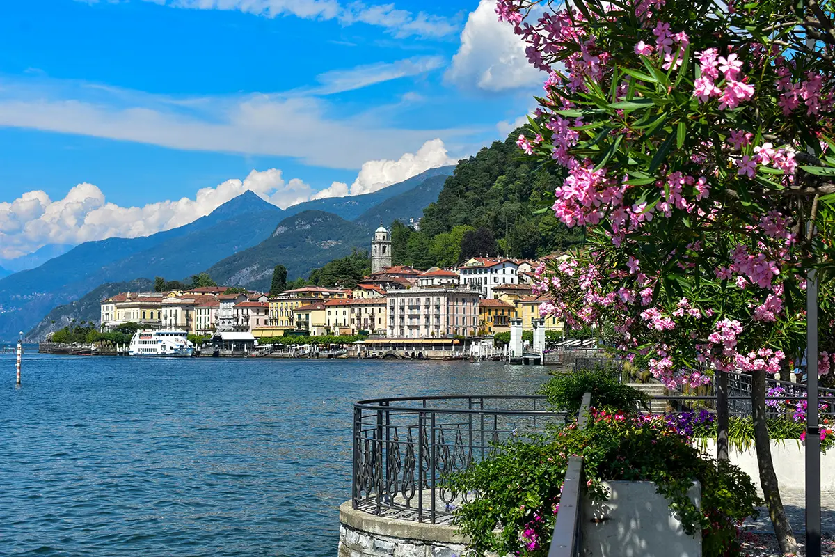 Pink flowers bloom along a stone railing, overlooking the blue waters of Lake Como and a picturesque town with mountain peaks in the background.