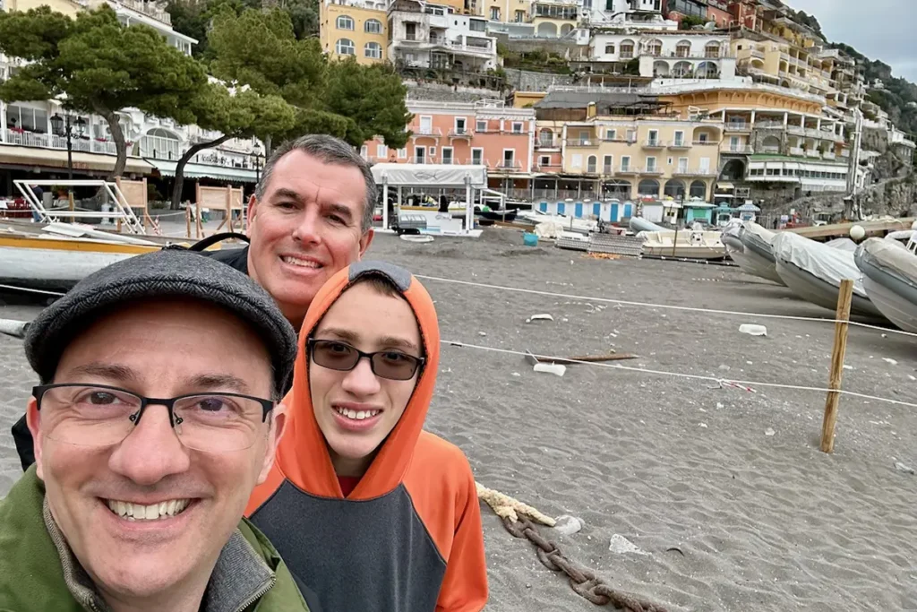 A selfie of a family standing on the beach in Positano, Italy, with colorful, tiered buildings in the background