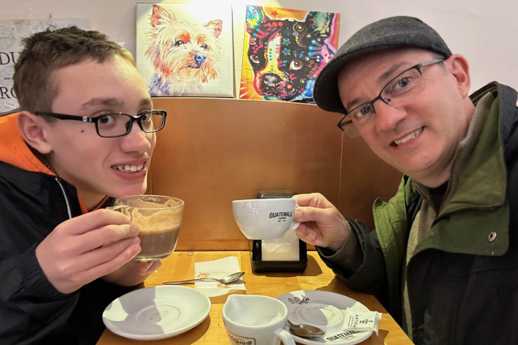 A man and his young son sit at a cafe table, smiling and holding up their coffee cups as if toasting the camera.
