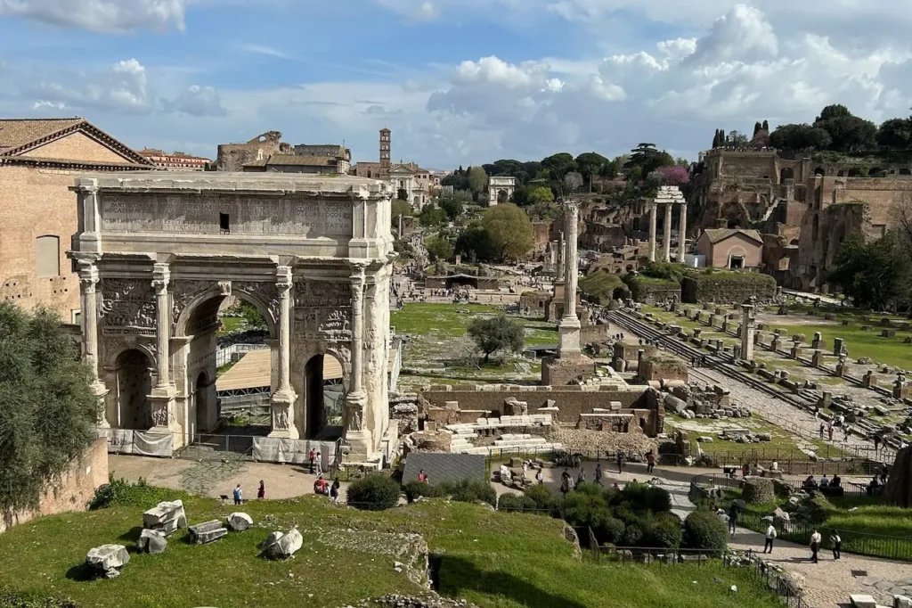 Panoramic view of the ancient Roman Forum ruins under a clear blue sky, featuring stone columns, arches, and the Temple of Saturn.