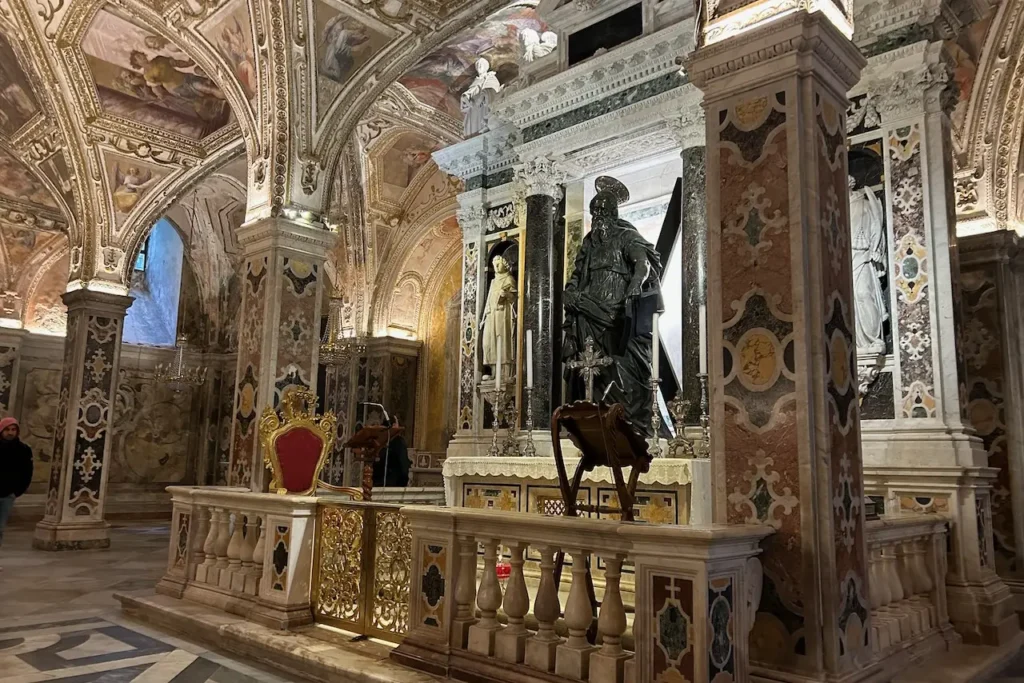 The ornate interior of the Amalfi Cathedral features marble pillars, vaulted frescoed ceilings, and a large bronze statue behind a central altar.