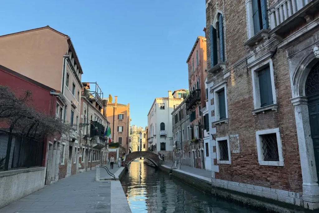 A narrow canal in Venice's Dorsoduro district is lined with historic brick buildings and a small arched bridge under a clear blue sky.