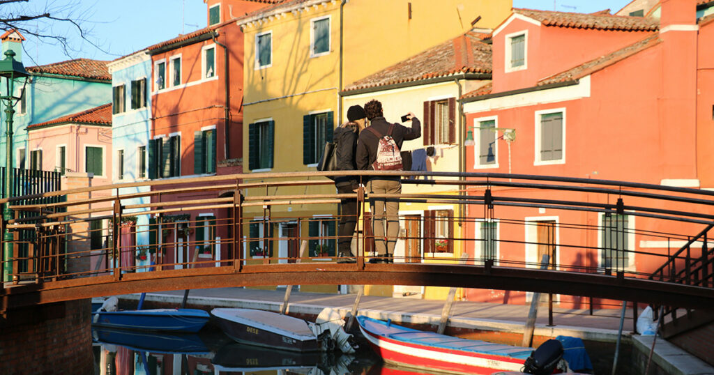 Two people stand on a small bridge in Burano, Italy, taking a photo of the vibrant yellow, orange, and red buildings reflected in the calm canal water below.