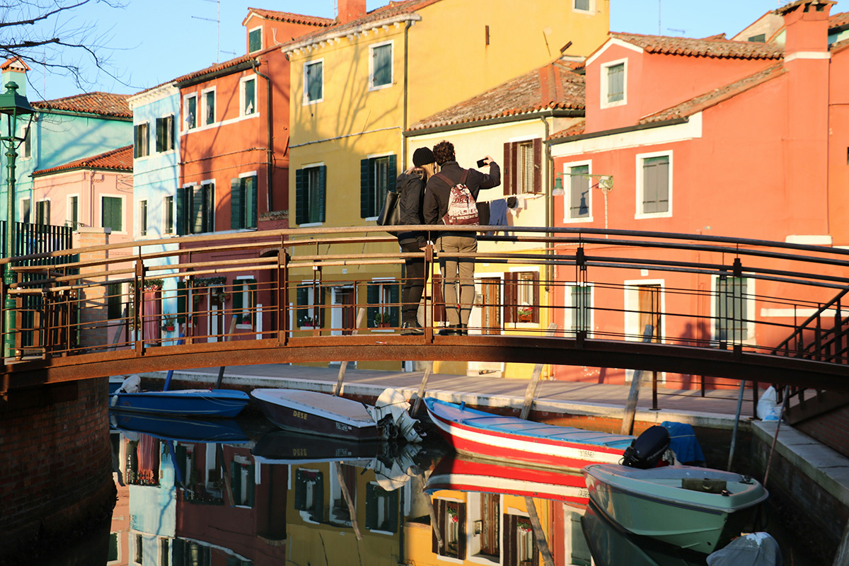 A couple stands on a small bridge in Burano, Italy, taking a photo of the vibrant yellow, orange, and red buildings reflected in the calm canal water below.