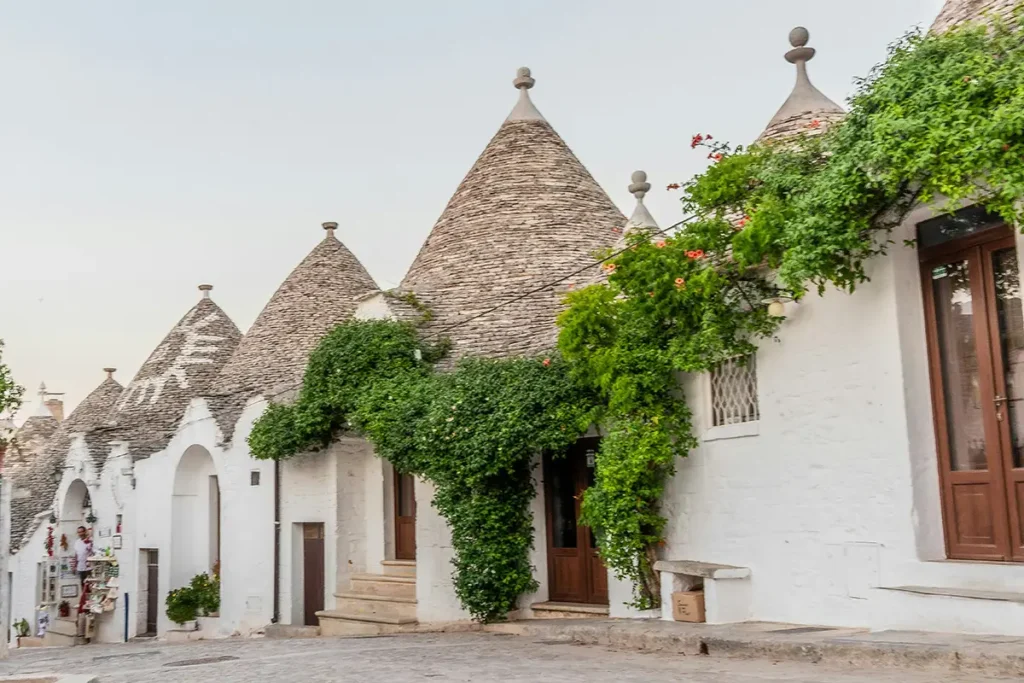 A row of traditional white trulli houses in Alberobello, Italy, featuring conical stone roofs and green vines under a pale sky.