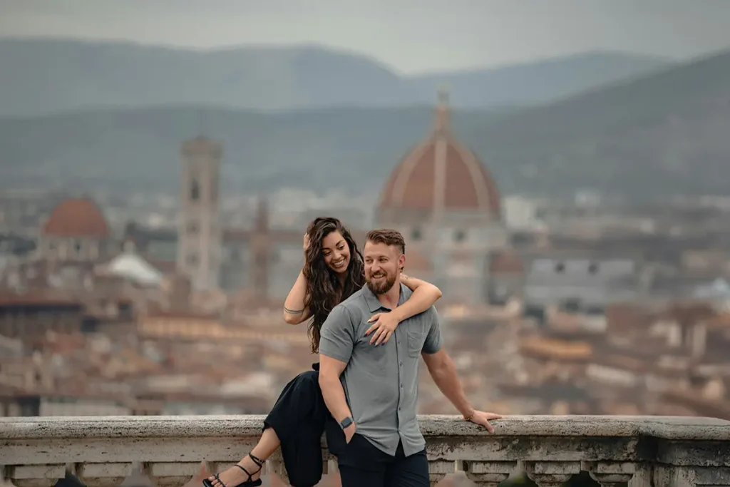 A smiling couple poses on a stone terrace overlooking the city of Florence, with the iconic Brunelleschi's Dome visible in the blurred background.