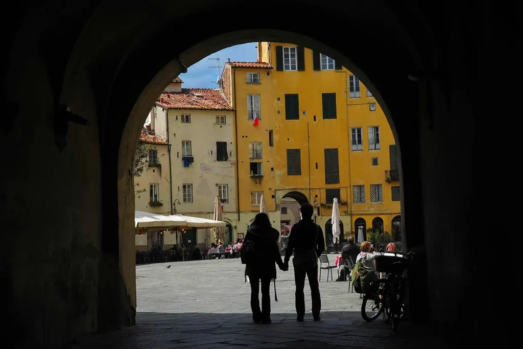A couple holds hands while walking through a dark stone archway into the sunlit, yellow-walled Piazza dell'Anfiteatro in Lucca, Italy