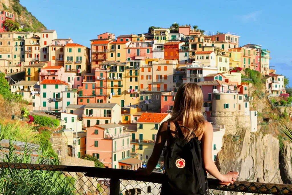 A young woman with a black backpack overlooks the colorful, cliffside buildings of Manarola, Italy, from a wooden railing under a bright blue sky.