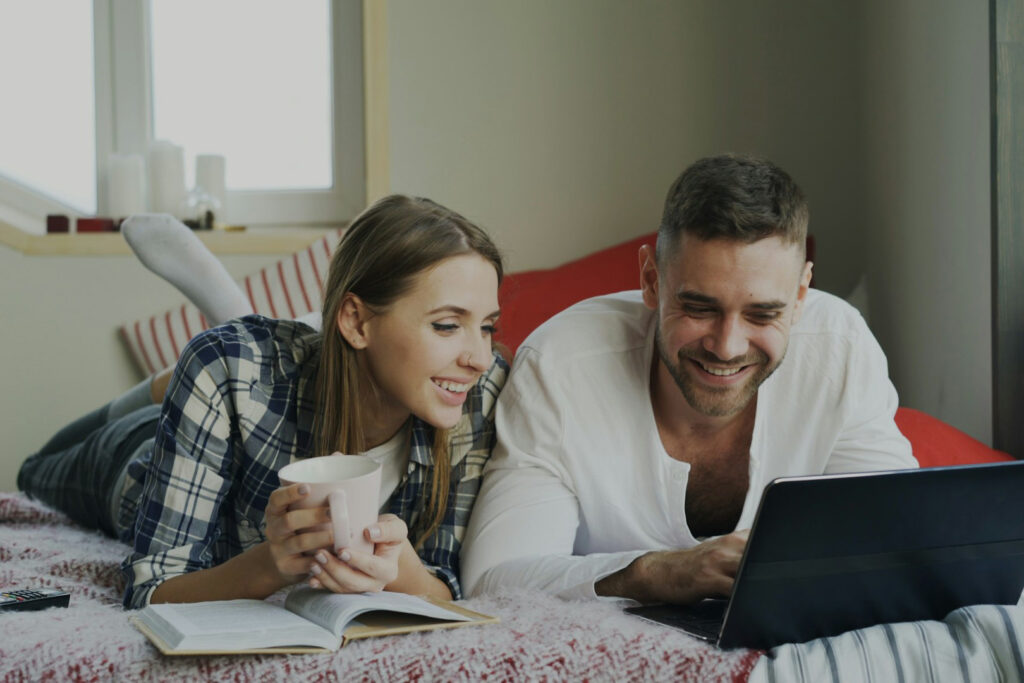 A couple is laying on a bed looking at a computer screen. The woman has a book open in front of her and is holding a mug.