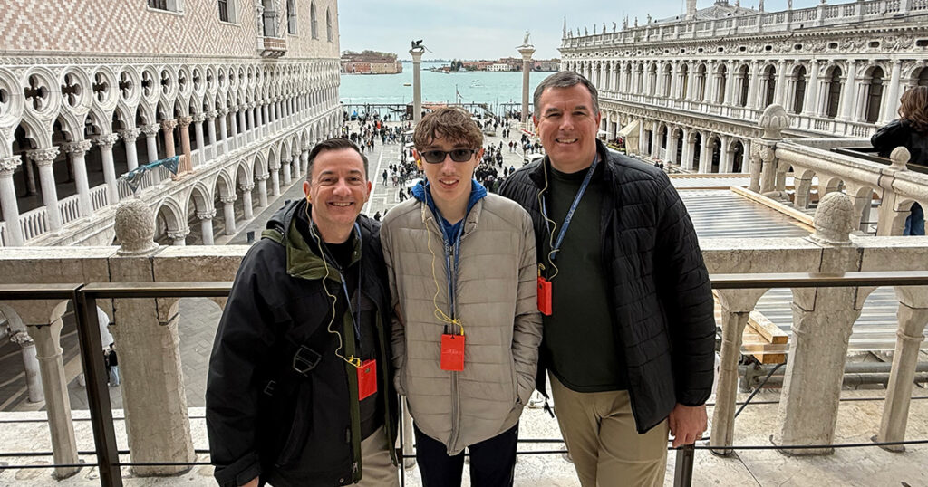 In this Italy trip with teenagers, a family of three smiles for a photo on a St. Mark's Square balcony in Venice.