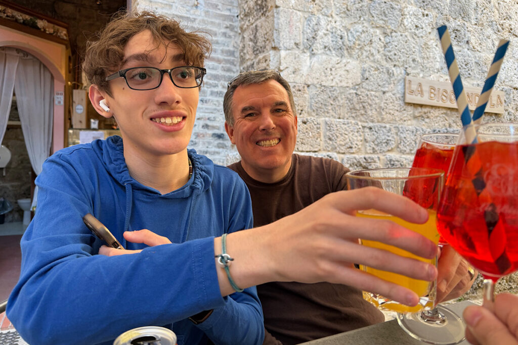 A smiling man and teenager toast with drinks, enjoying the aperitivo tradition at an outdoor cafe against a rustic stone wall in Italy.