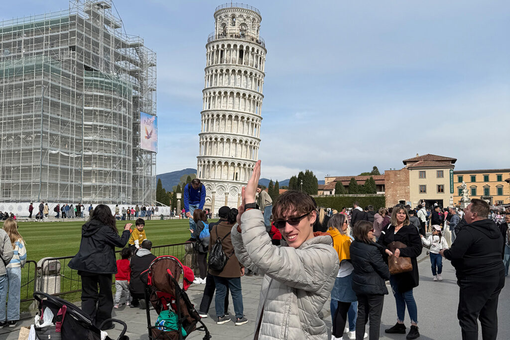 A young man in a gray jacket poses in front of the Leaning Tower of Pisa, holding his hands up to appear as if he is supporting the tilting structure.
