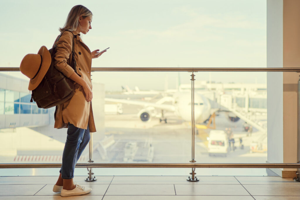 A woman wearing a trench coat and a backpack looks at her phone in an airport terminal, with an airplane visible out the window.
