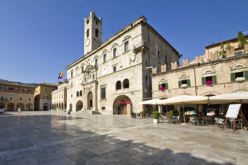 The historic Palazzo dei Capitani with its tall clock tower stands in the wide, sunlit Piazza del Popolo in Ascoli Piceno, Italy.