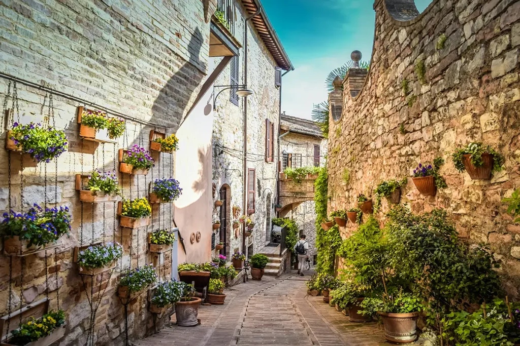 A narrow stone alley in Spello, Italy, lined with historic brick houses decorated with colorful potted flowers and green vines.