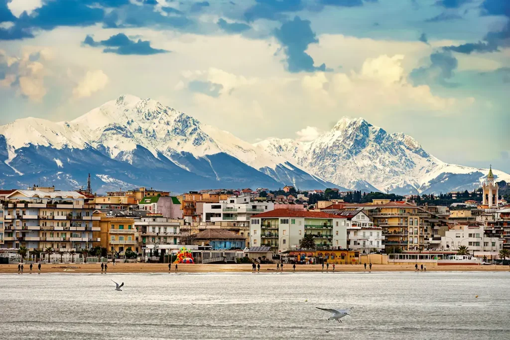 The coastal city of Pescara, Italy, sits along a sandy beach with seagulls flying over the water, backed by massive, snow-covered mountains.