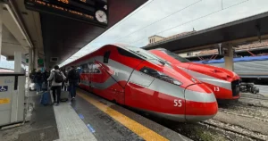 Passengers walk past a red Frecciarossa high-speed train at an Italian railway platform under an electronic departure sign showing Napoli.