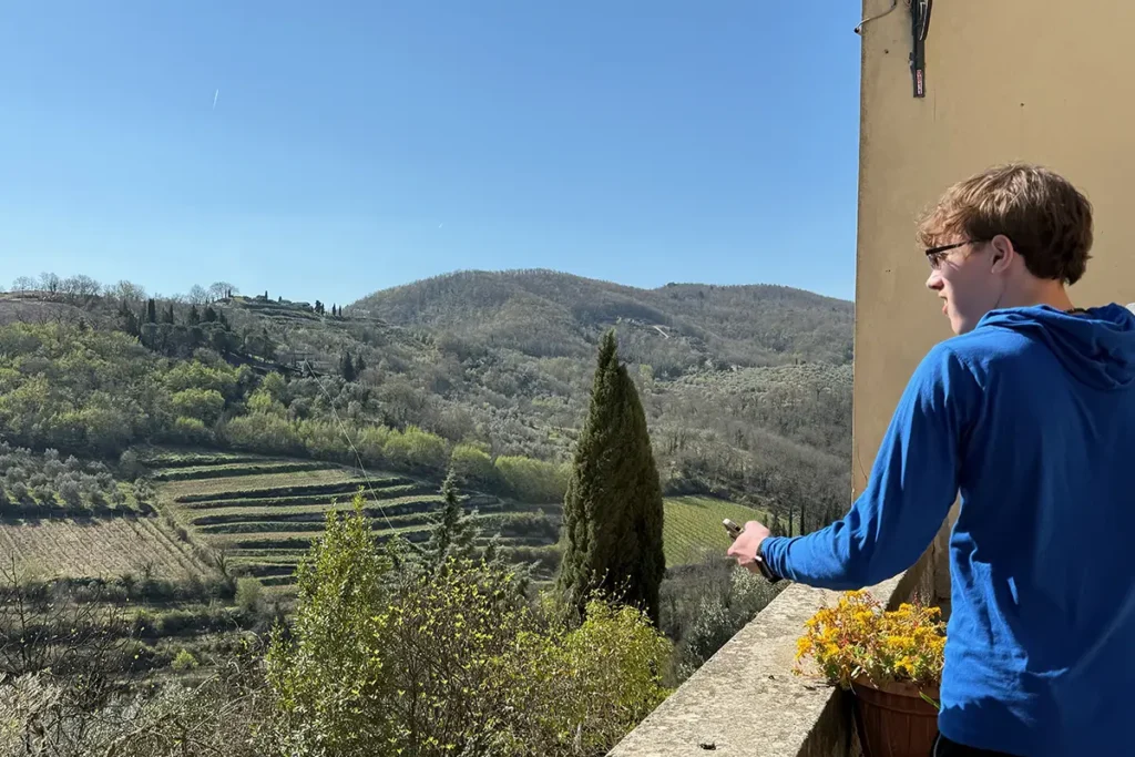 A person in a blue hoodie looks out from a balcony over a sunlit valley of terraced hills and forests in Montefioralle, Italy.