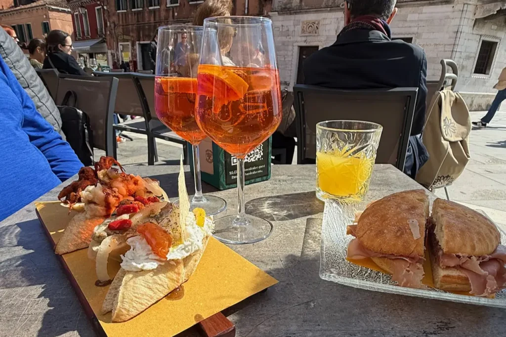 An outdoor table in Venice, Italy, set with Spritz cocktails, sandwiches, and a board of assorted Venetian cicchetti appetizers.