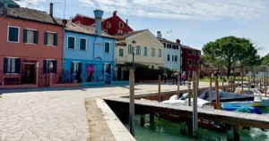Vibrant, colorful houses line a paved waterfront promenade where boats are docked along a wooden pier in Burano, Italy.