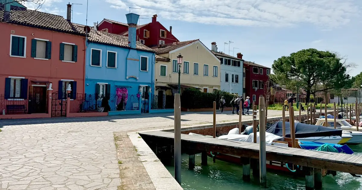 Vibrant, colorful houses line a paved waterfront promenade where boats are docked along a wooden pier in Burano, Italy.