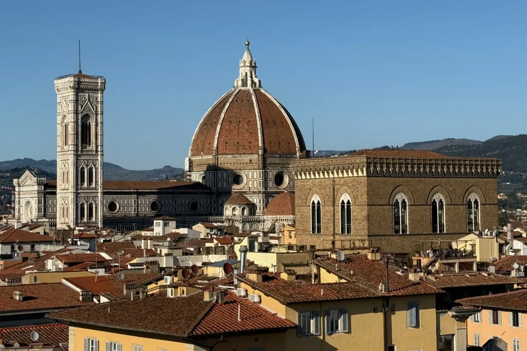 The iconic Duomo dome and Giotto's Campanile rise above the red-tiled rooftops of Florence, Italy, against rolling hills and a clear blue sky.